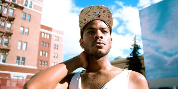 A young black man in a white tank and wearing a a yellow patterned baseball cap stands outside in a city on a sunny day. He has one arm on his neck and