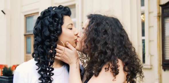 Two young women with long curly hair kiss outside on a city street.