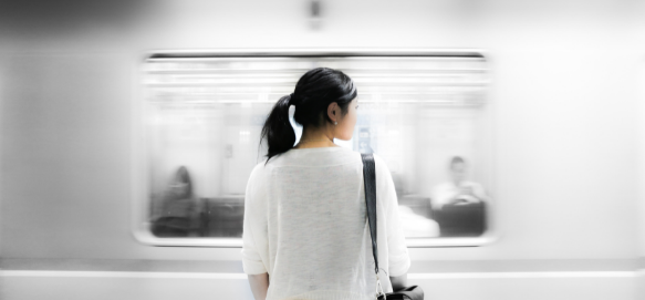 A woman stands on a subway platform looking away from the camera as a train speeds by.
